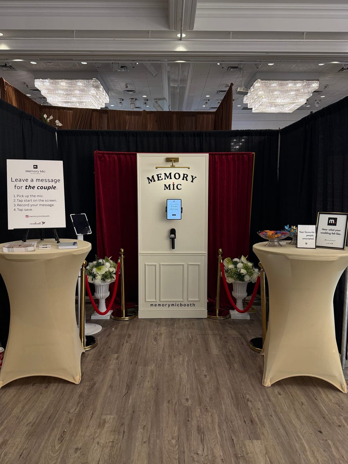 Memory Mic video guestbook booth at the South Asian Bride show: an ivory panelled booth with the couple's names on the screen, a branded microphone, red velvet backdrop, gold stanchions and white florals.